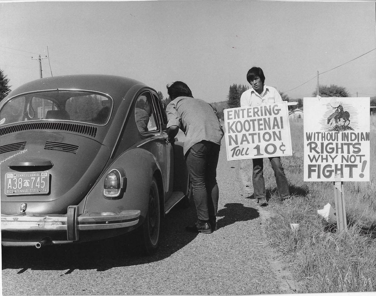 Two young Kootenai men beside a Volkswagen at a roadside toll stop in 1974, with signs reading Entering Kootenai Nation, toll 10 cents
