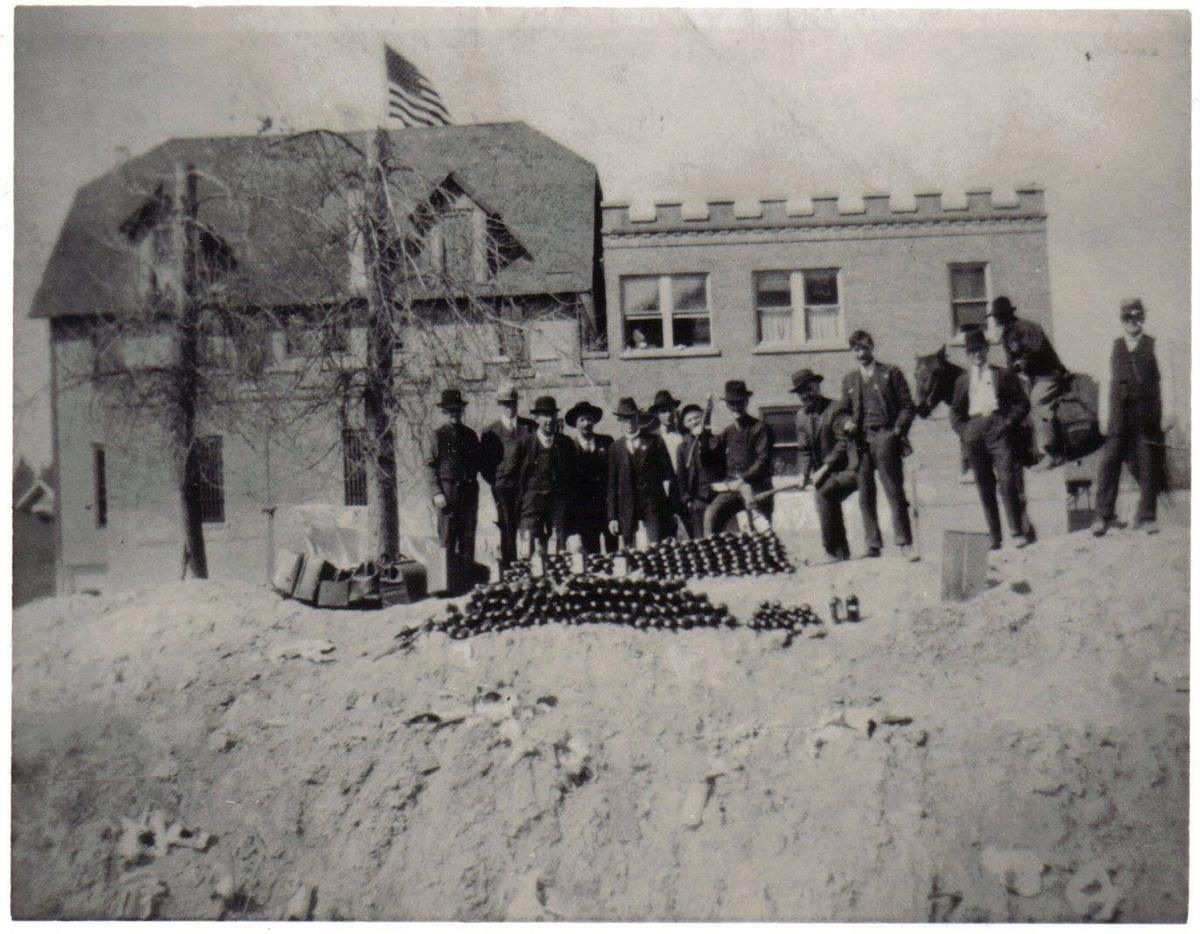 A group of men in hats stand behind rows of confiscated bottles lined up on the ground in front of a brick building, Prohibition era
