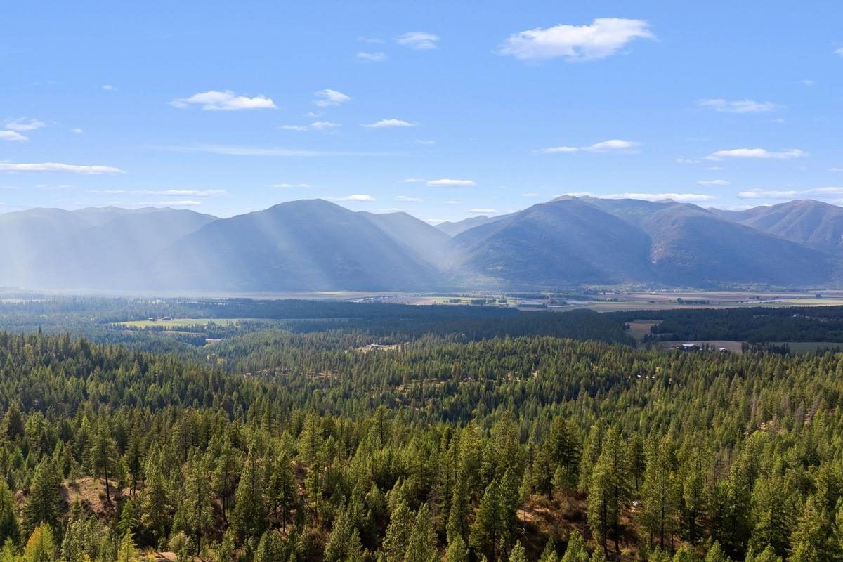 Wide view across timbered benches and the Kootenai Valley to the Selkirk Mountains, with sun rays breaking through the clouds