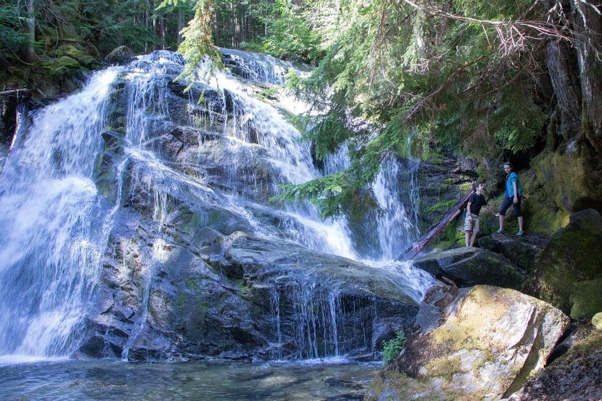 A wide waterfall cascading over mossy granite into a clear pool, with two hikers standing on the rocks beside it