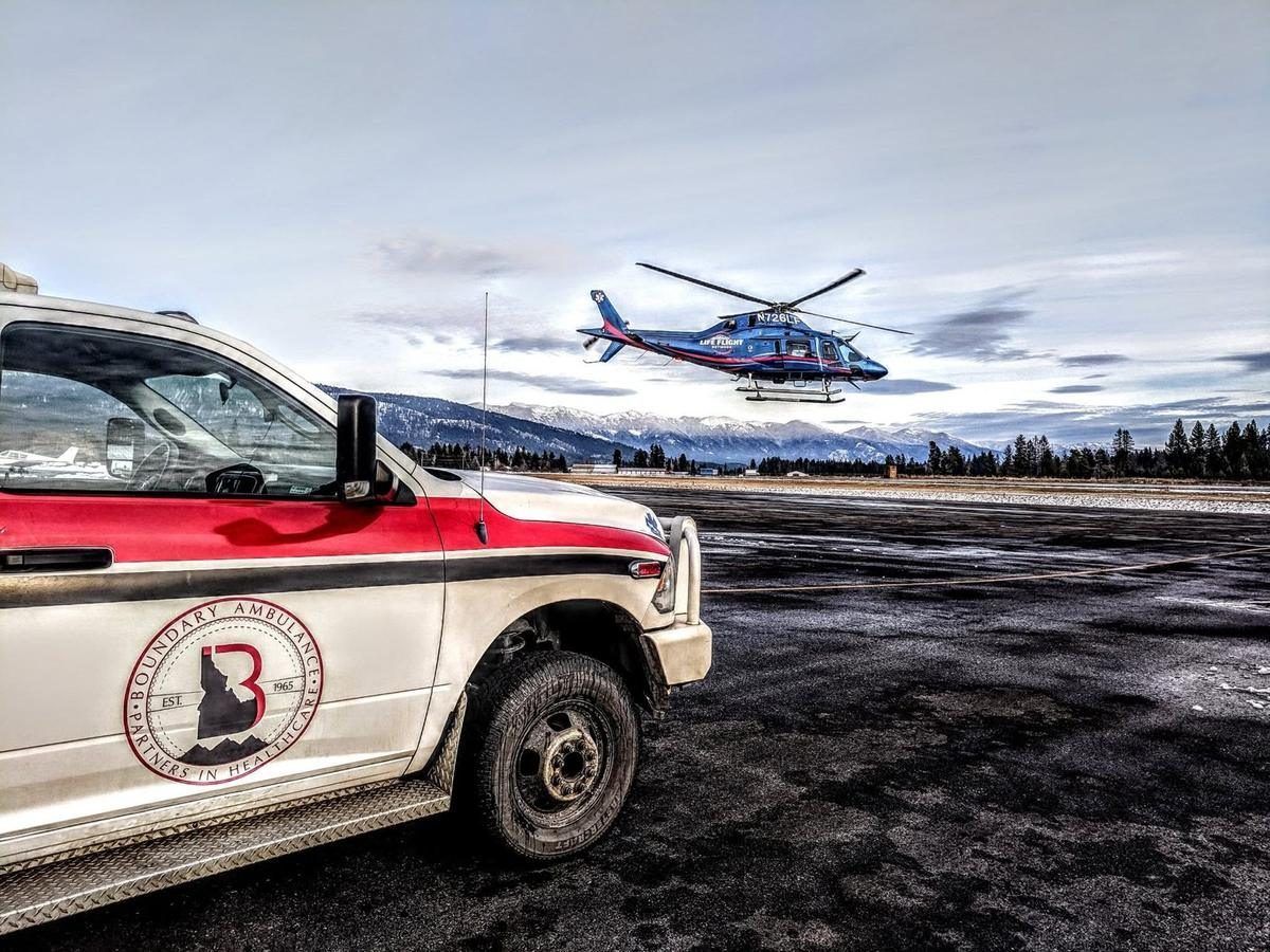 A Boundary Ambulance vehicle on the tarmac as a Life Flight Network helicopter lifts off, with the snow-capped Selkirk Mountains behind