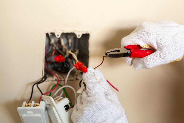 Gloved hands handling red electrical wires inside an open wall outlet box