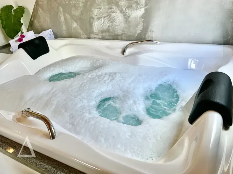 Close de uma banheira de hidromassagem branca com espuma e água em movimento, travesseiros de apoio pretos e parede cinza marmorizada ao fundo na Quattuor Cabanas em São Miguel do Gostoso.