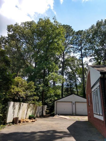 Driveway beside a brick house, with a detached garage, tall trees, and a white fence in bright daylight