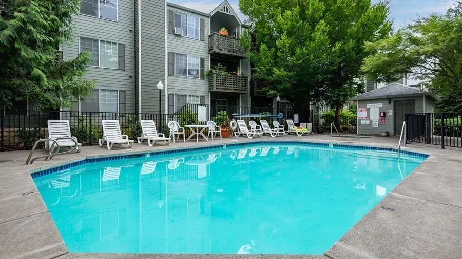 Swimming pool in front of apartment building, with lounge chairs and trees around it.