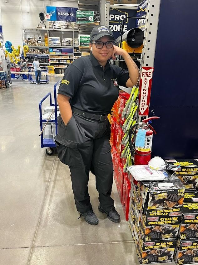 Person in a Lowe's store uniform leans against a display, resting hand on face. A fire extinguisher and merchandise are nearby.