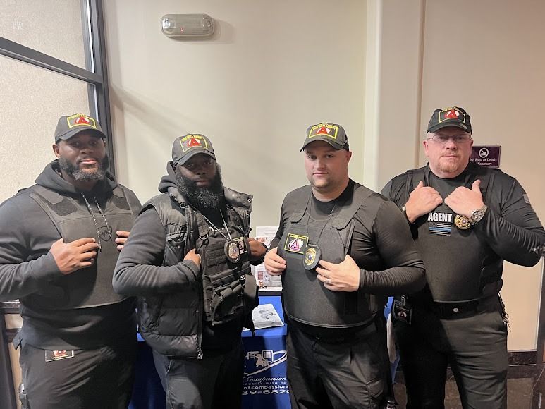 Four people in black uniforms and vests, standing indoors near a table, some touching their vests.