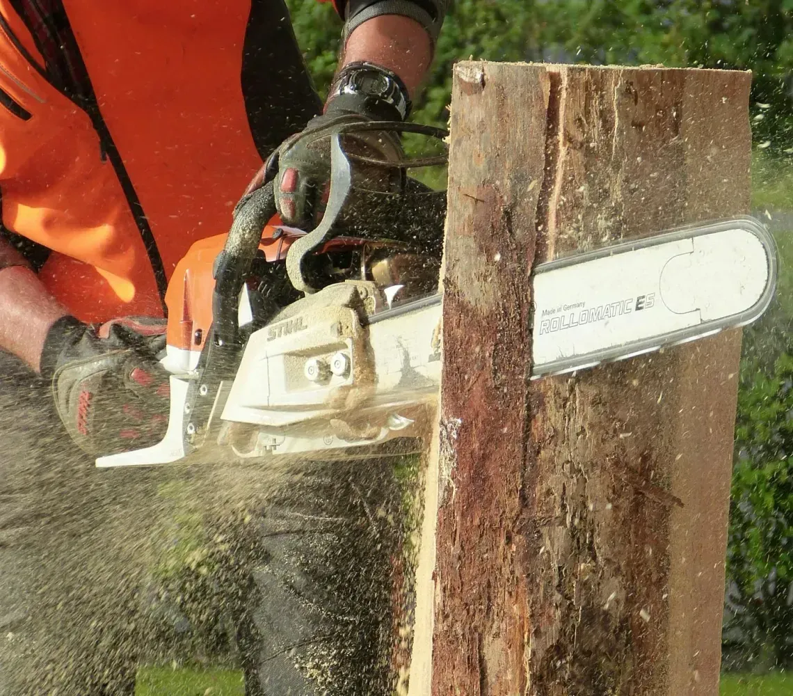 Person cutting a tree trunk with a chainsaw, wood chips flying, outdoors.