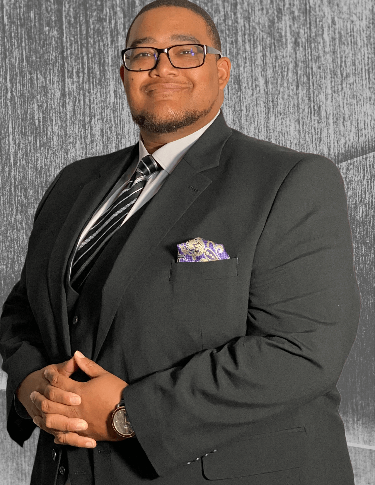 Man in suit with glasses, hands clasped, smiling