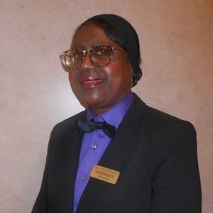 Woman in black blazer, purple shirt, and bow tie, wearing glasses, name tag visible.