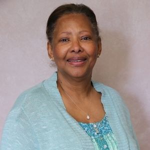 Woman smiling, wearing a blue sweater and necklace, against a light background.