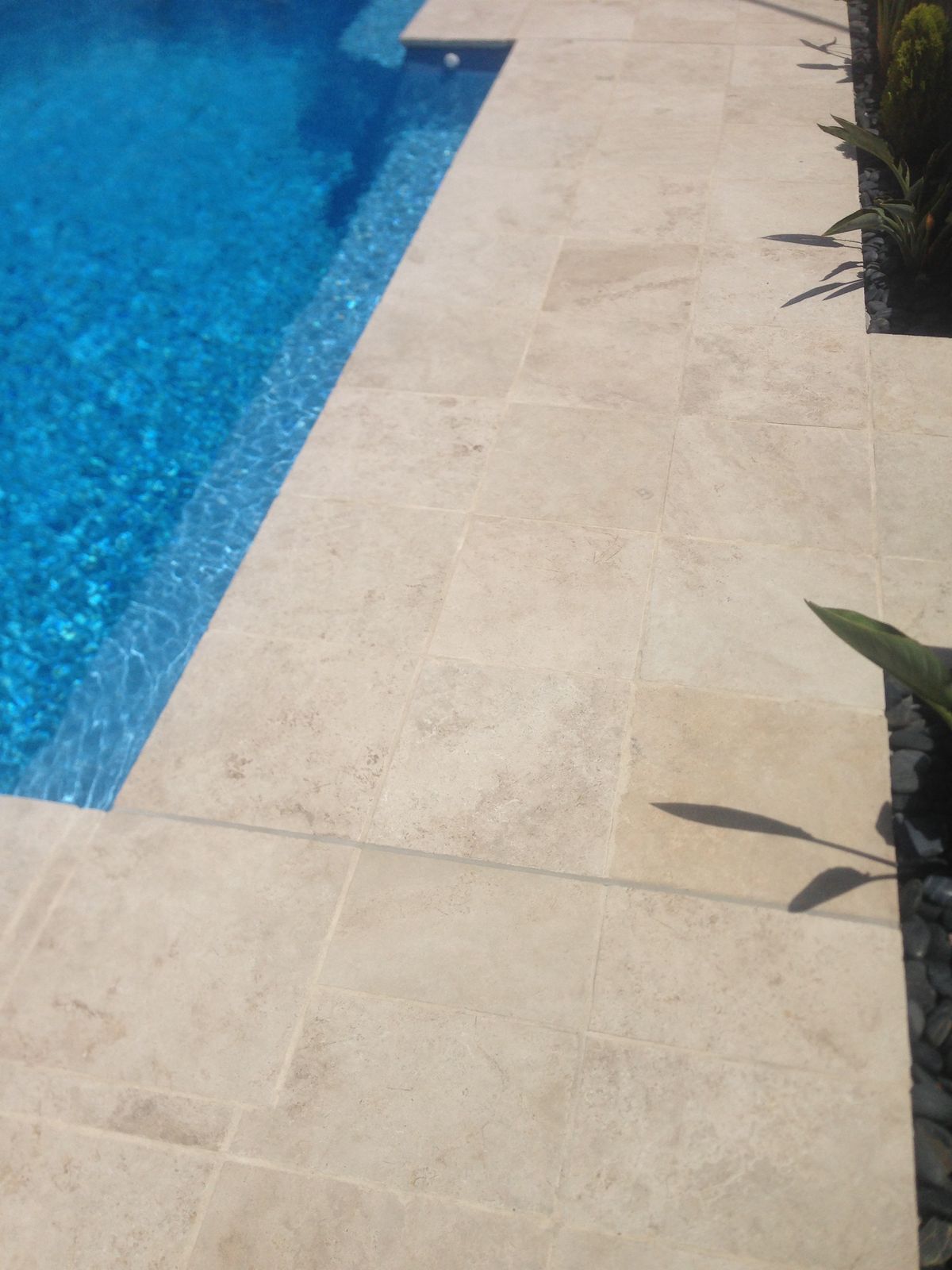 Beige stone pool deck beside bright blue swimming pool water, with plant shadows on the right.