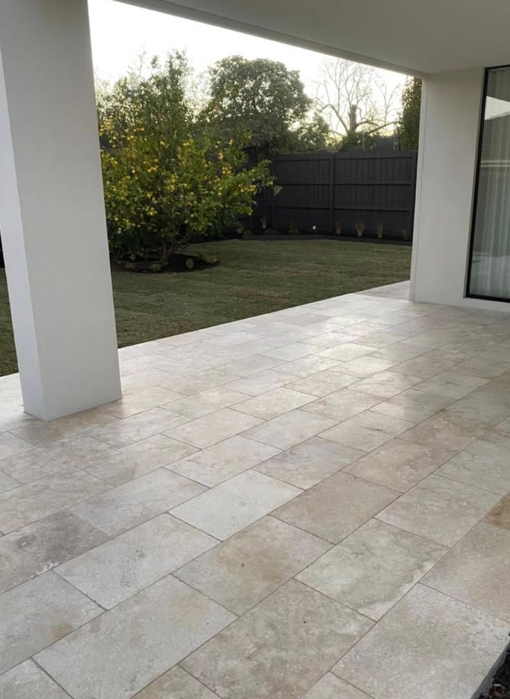 Covered patio with light stone tiles, white columns, and a fenced backyard with grass and trees.