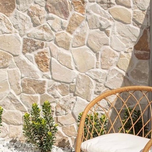 Stone wall with a wicker chair and white cushion beside small green shrubs outdoors
