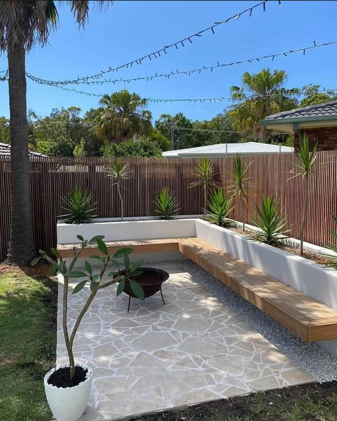 Small patio with white bench seating, potted plant, tiled floor, and string lights under a blue sky