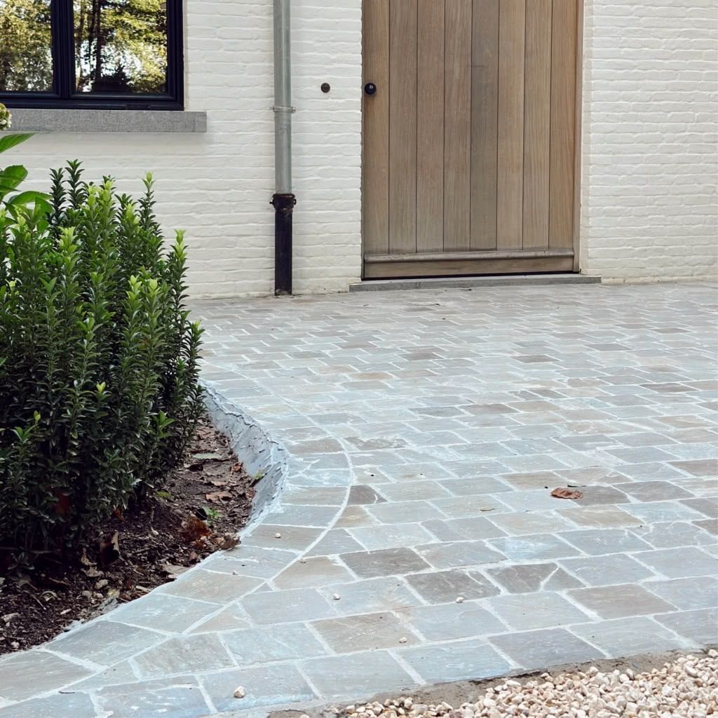 Curved stone walkway leading to a wooden front door beside a house window and shrubbery
