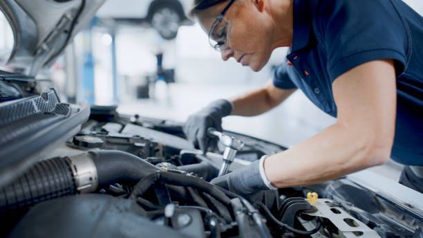 Mechanic wearing safety glasses and gloves works on a car engine in a shop.