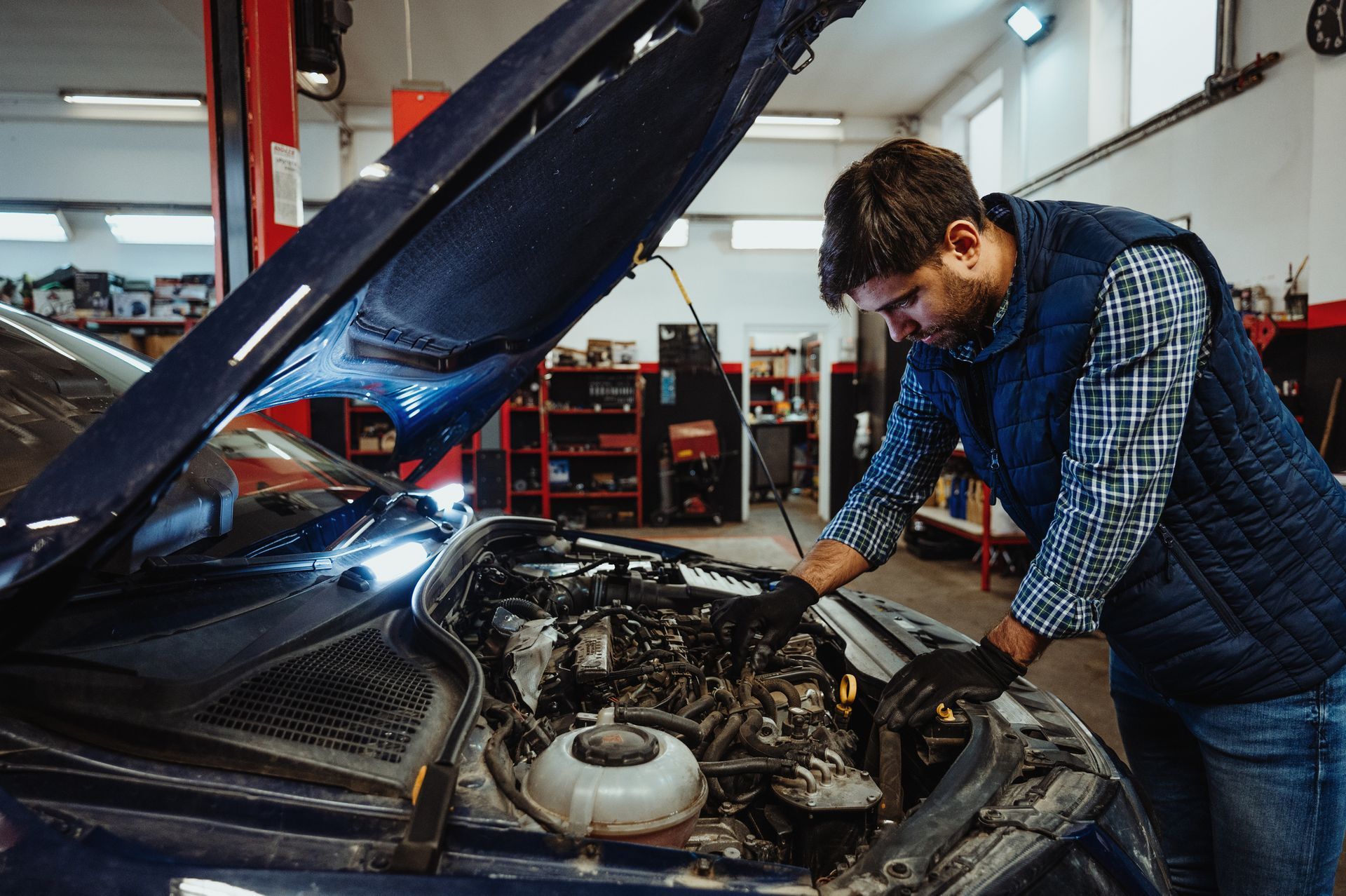 Mechanic in a blue vest, inspecting a car engine in a garage.
