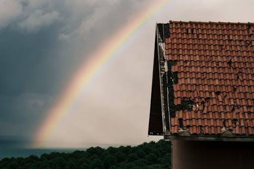 Residential tiled roof with visible storm damage and missing shingles under a rainbow sky.
