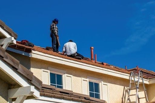 Roofing contractors installing and repairing roof shingles on a residential home with ladders and tools.