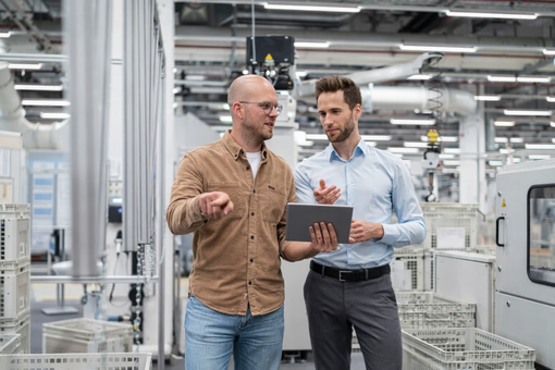 Twee professionals bespreken een tablet in een fabriekshal, met industriële machines op de achtergrond.