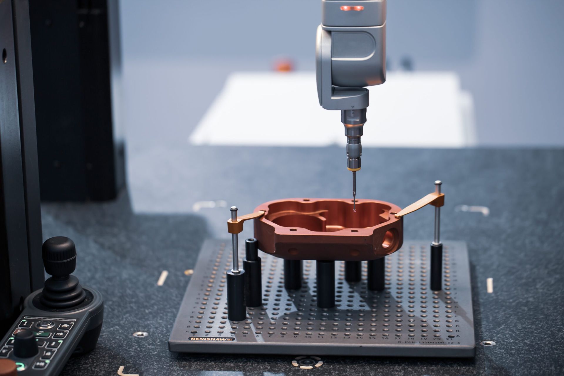 CNC-meetsonde meet een koperen onderdeel dat is vastgeklemd op een geperforeerde bevestigingsplaat, met een joystickcontroller in de buurt.
