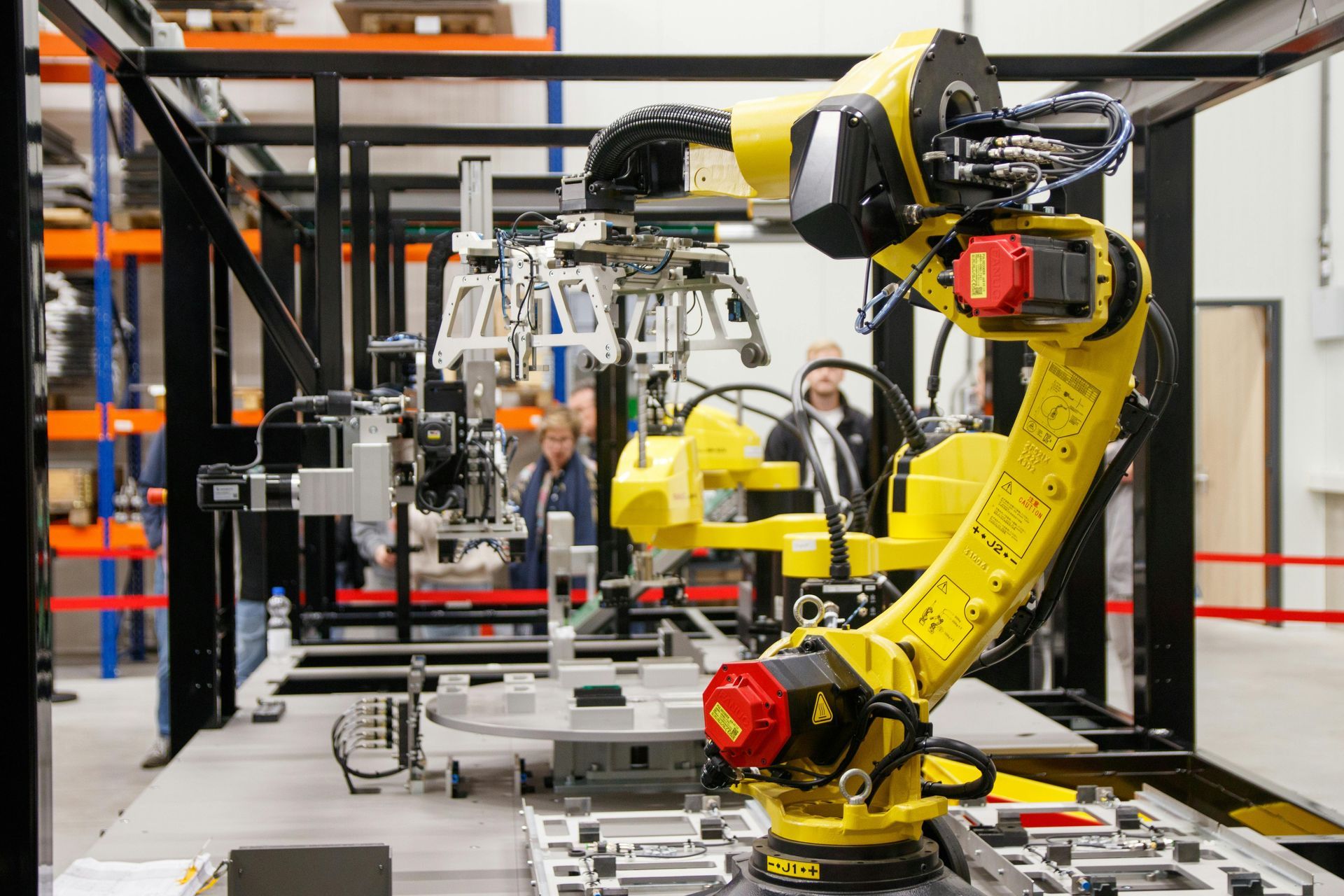 Een industriële robotarm assembleert onderdelen in een fabriek, met werknemers op de achtergrond.