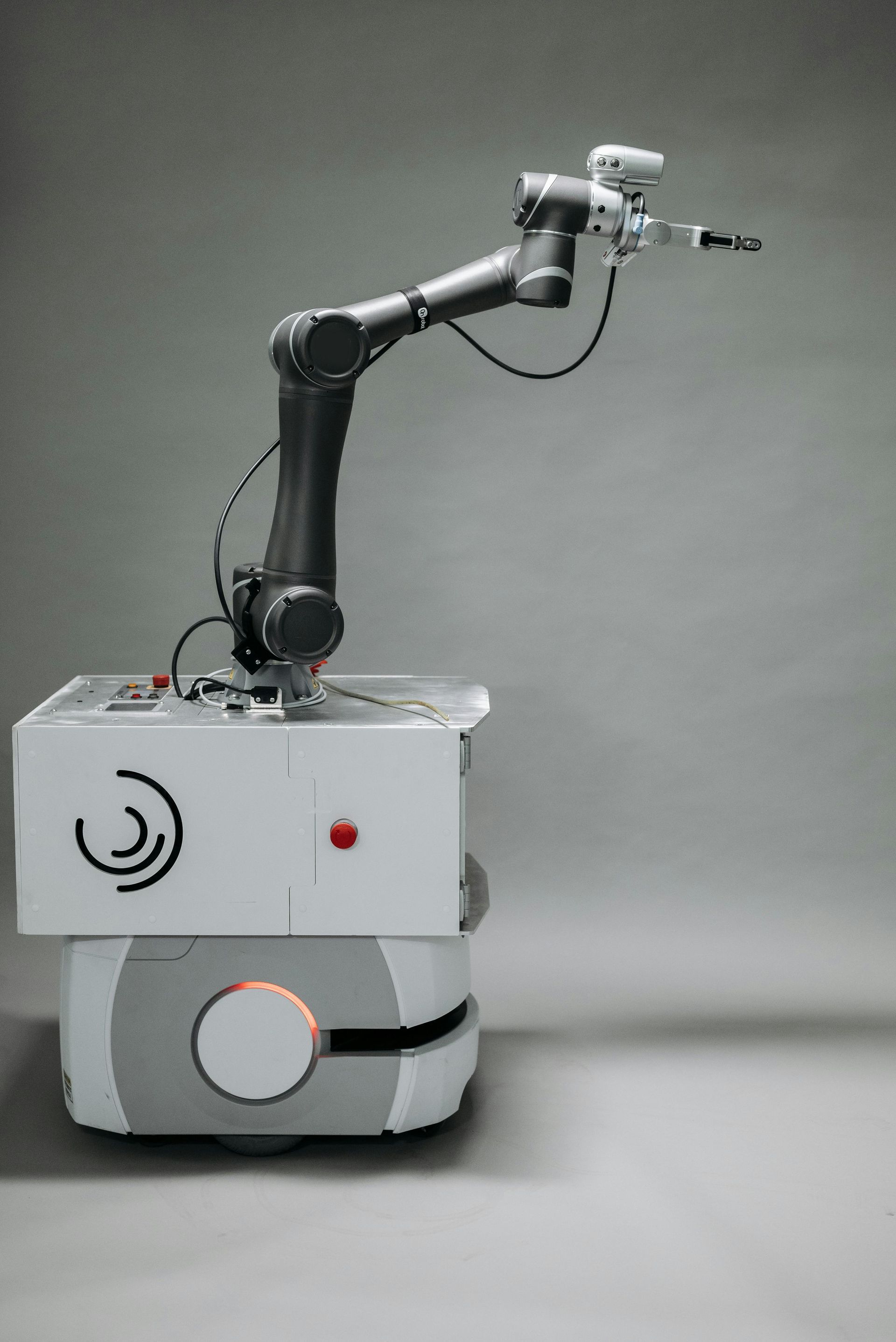 Kleine robot op wielen met een zwarte, beweegbare arm die een gereedschap vasthoudt, tegen een grijze achtergrond.