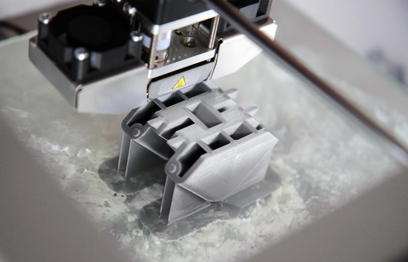 De nozzle van een 3D-printer print een klein grijs plastic onderdeel op de bouwplaat.