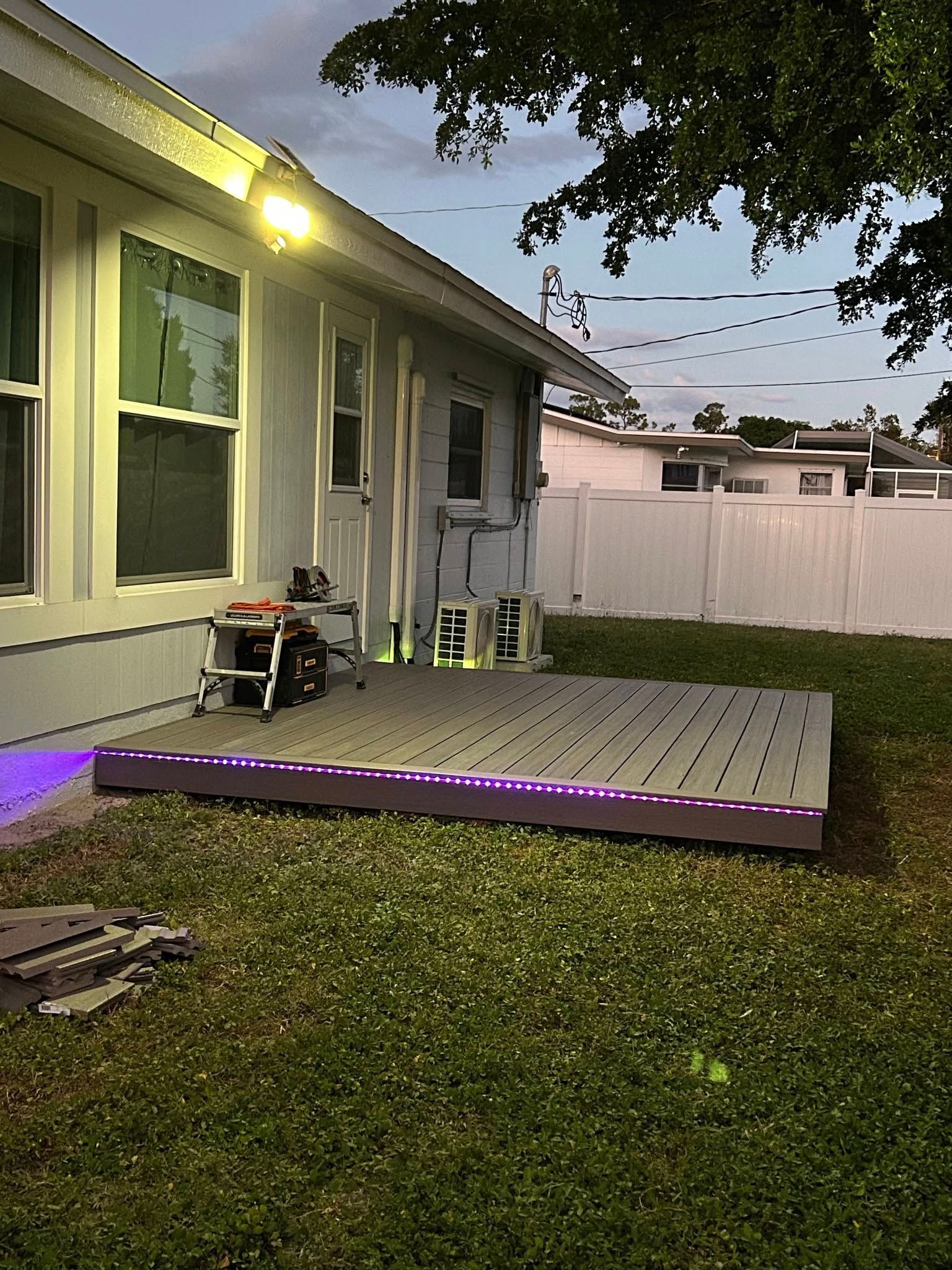 Backyard patio with lit house exterior, white fence, and a raised deck with purple lighting at dusk