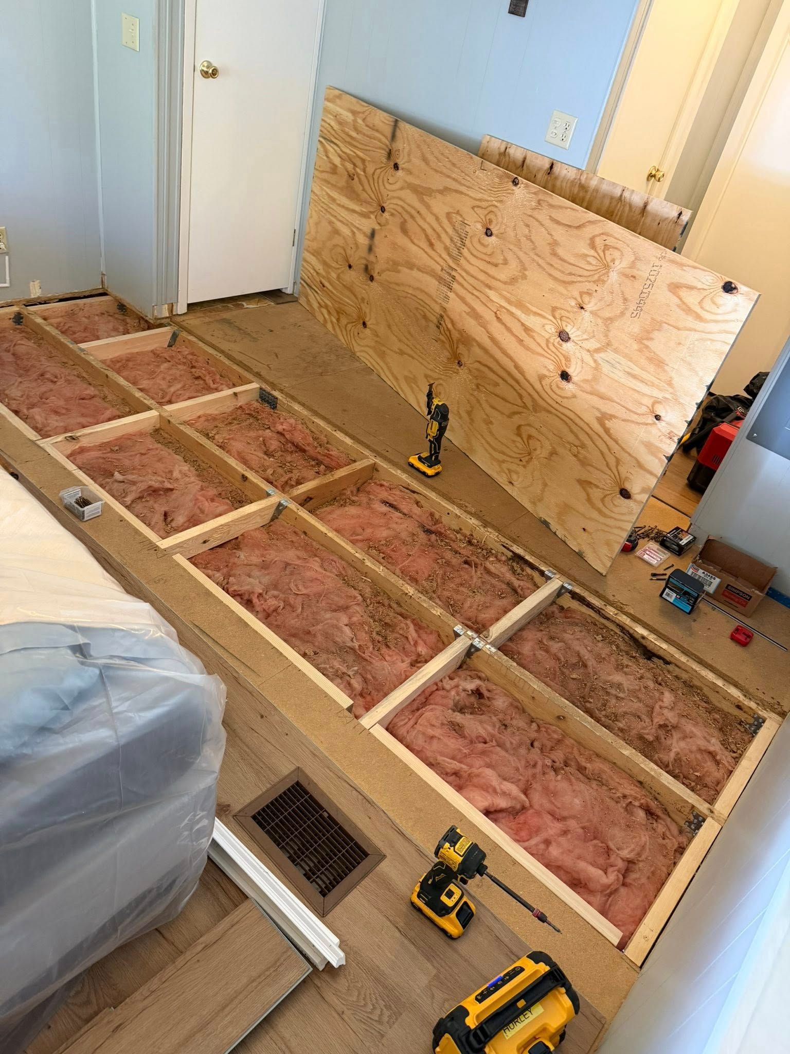 Partially gutted room with exposed subfloor, insulation, plywood wall cover, and tools on the floor