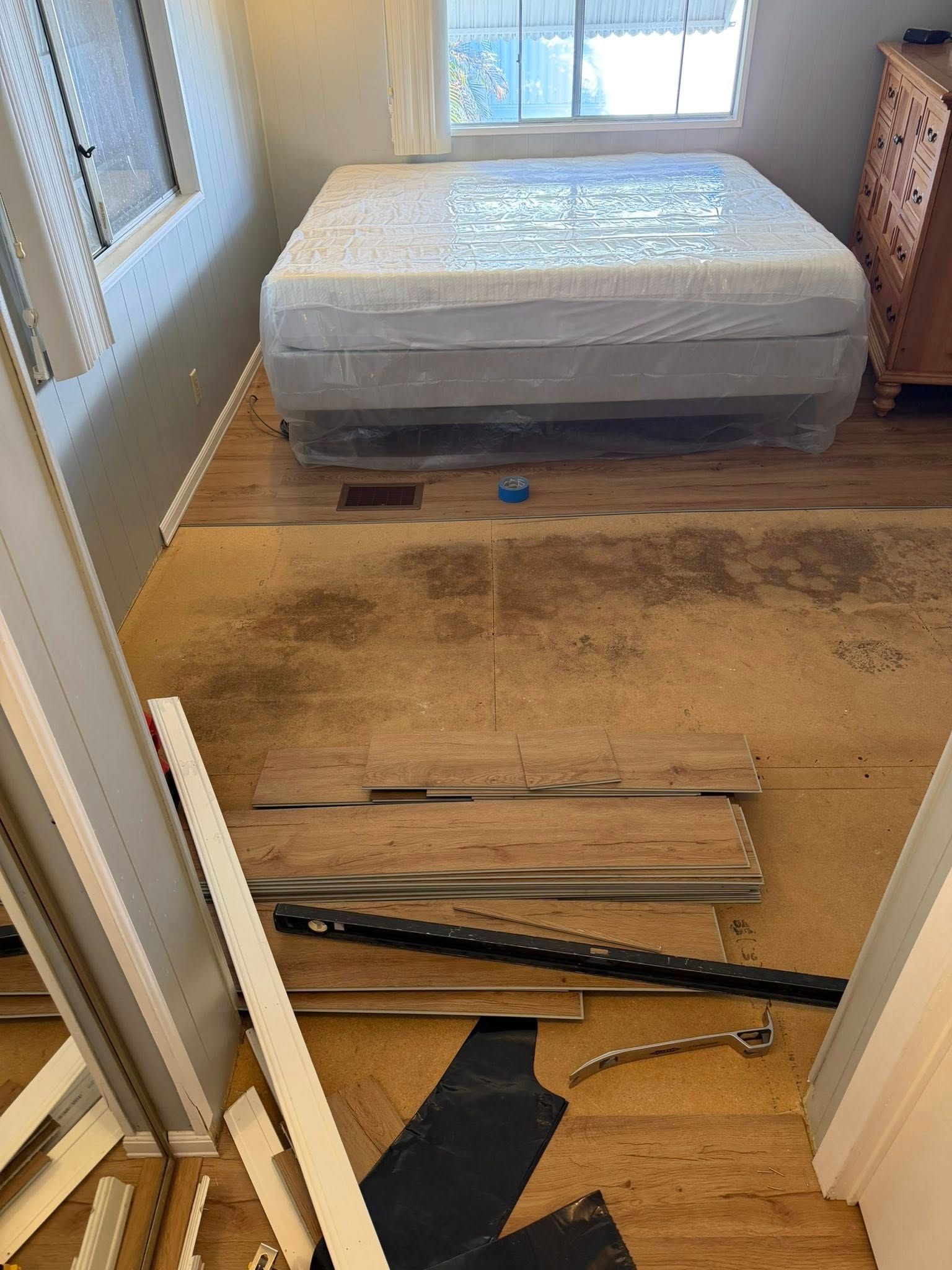 Messy bedroom with a wrapped mattress, scattered floorboards, and a glass door to the outside.