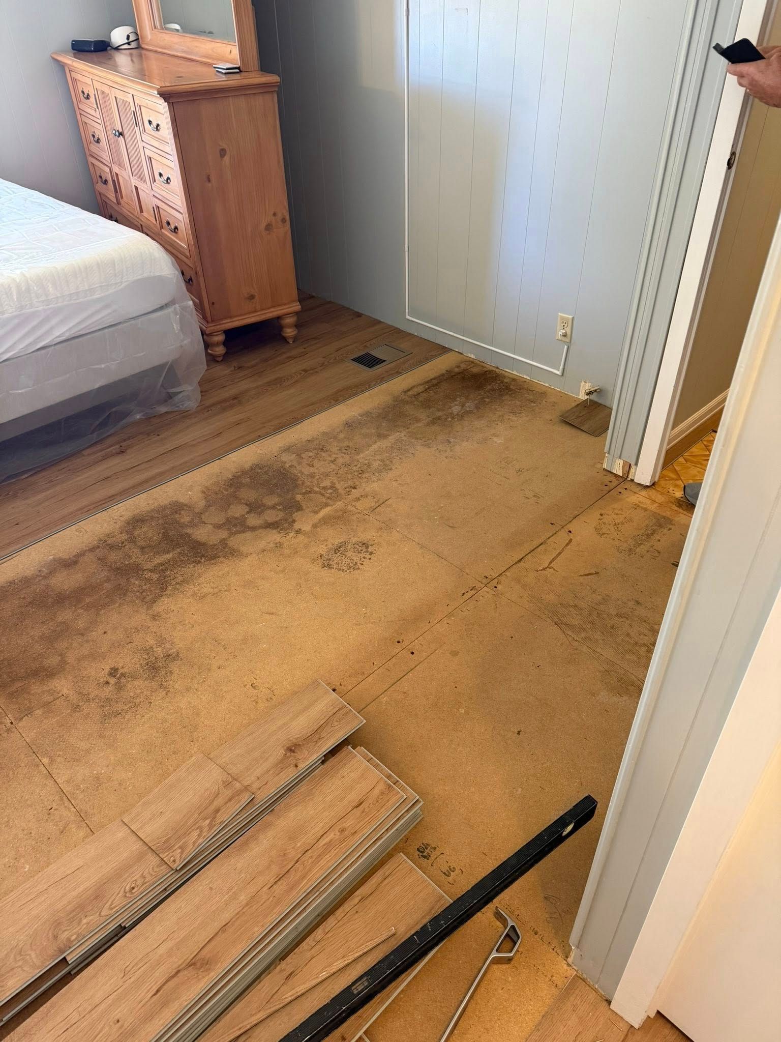 Damaged wooden floor with dark stains and broken boards in a bedroom near a bed and dresser.