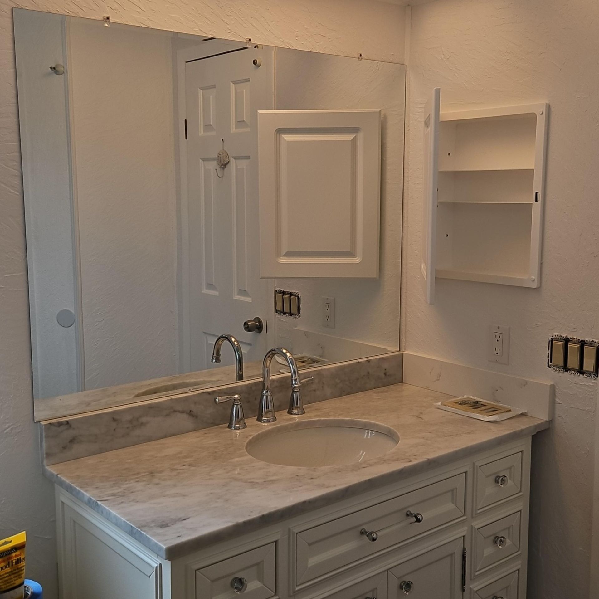 Bathroom vanity with mirror, marble countertop, sink, chrome faucet, and white wall shelf.