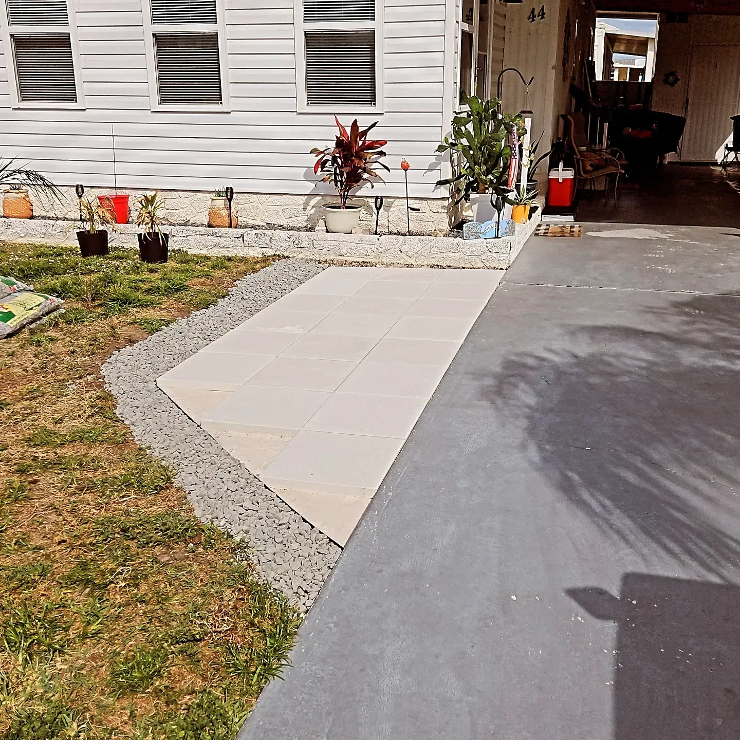 Walkway beside a house, with tiled path, gravel edging, grass, and potted plants near the entrance