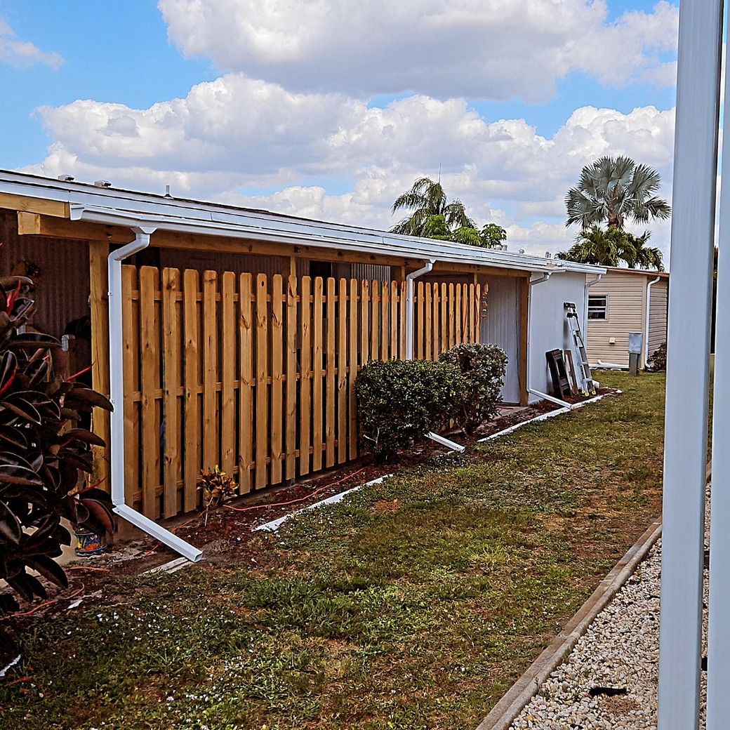 Wooden slatted fence beside a house with a grassy path, shrubs, and a partly cloudy sky.