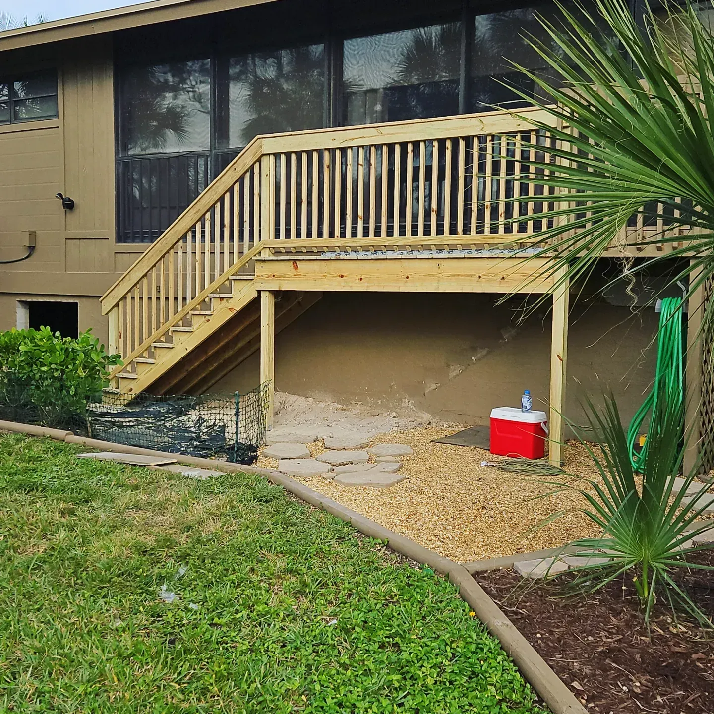 Wooden deck with stairs beside a beige apartment building, bordered by grass and landscaping.