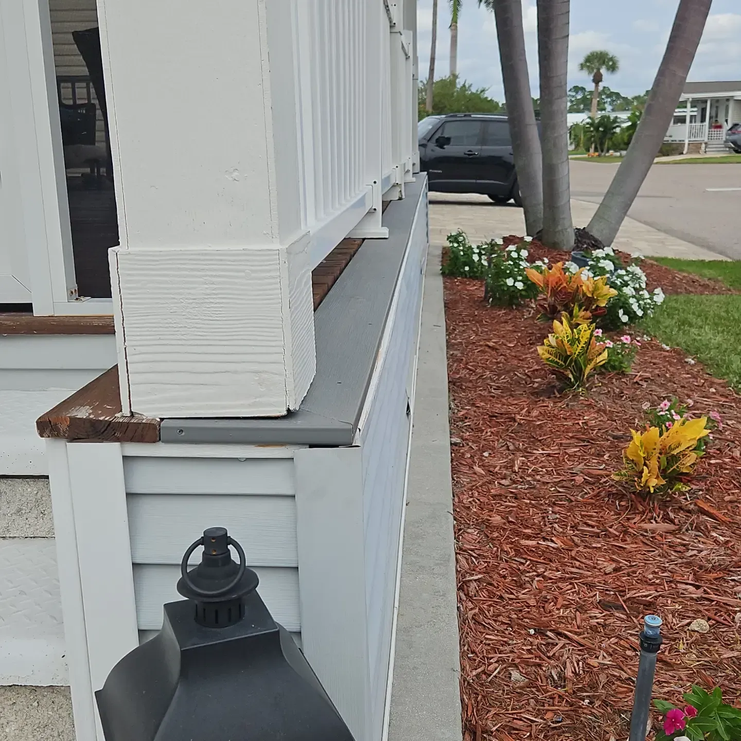 White porch corner with black lantern, beside a landscaped flower bed and sidewalk.