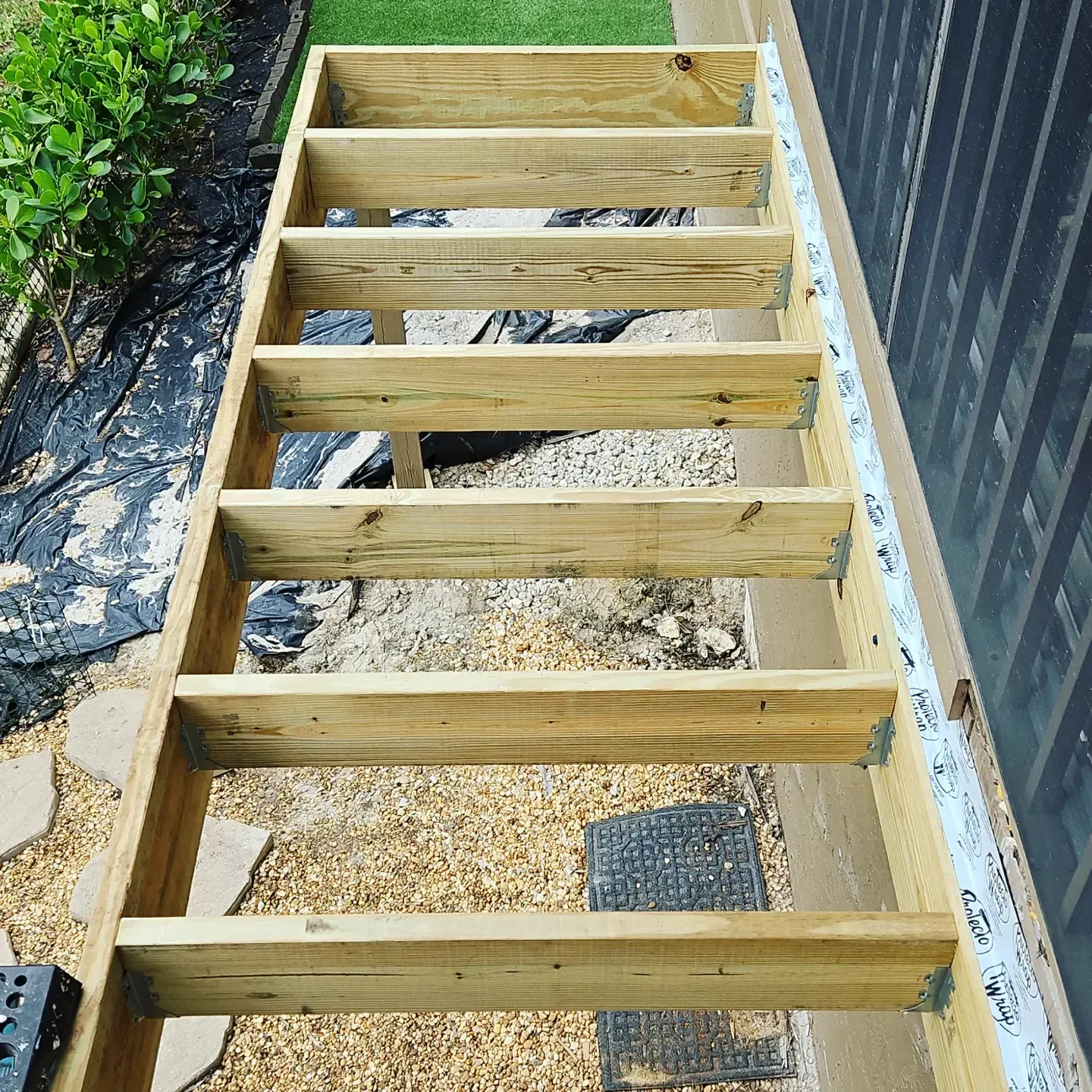 Wooden deck frame with evenly spaced joists over gravel beside a dark fence and greenery