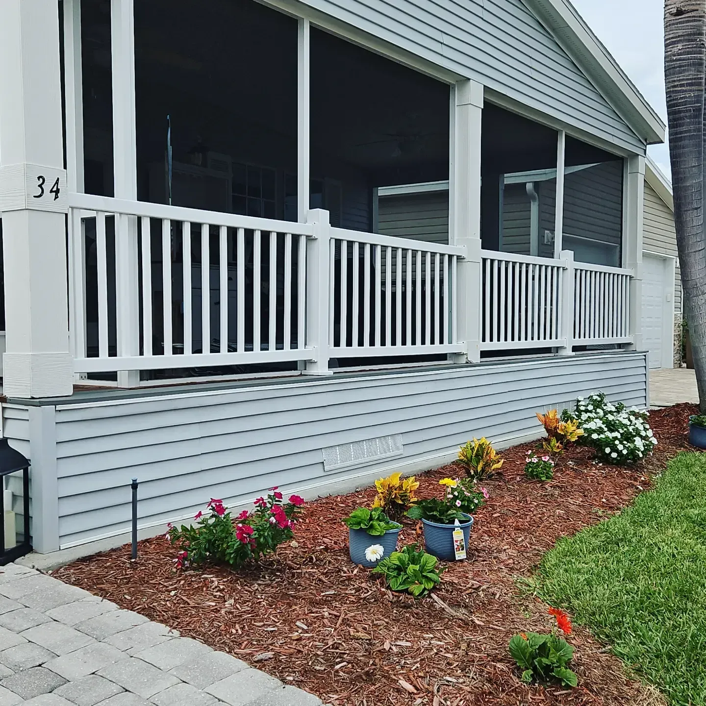 Front porch with white railing, gray siding, and a colorful flower bed beside a paved walkway