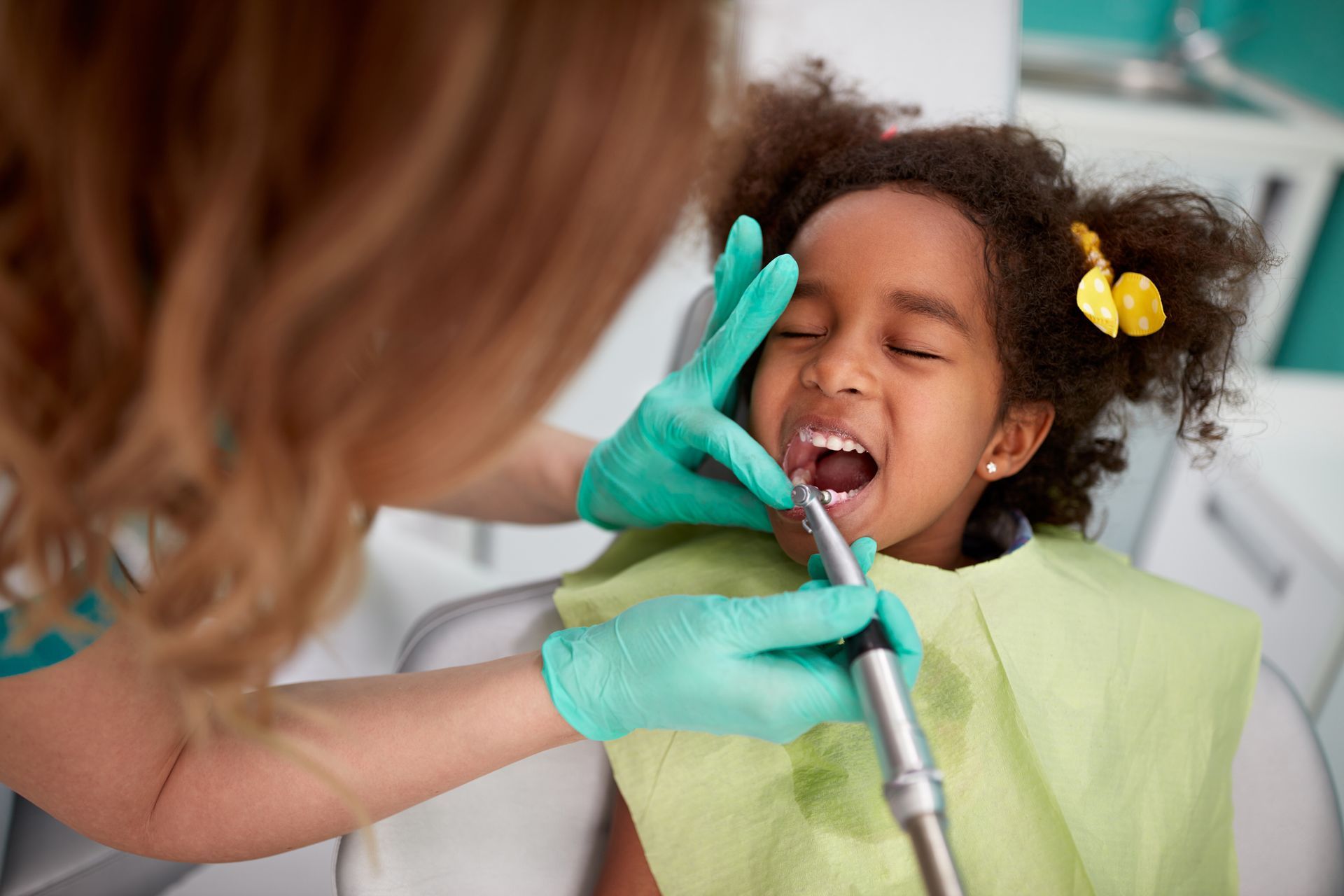 Dentist cleaning a child’s teeth with a dental drill while the child sits in a chair with a bib