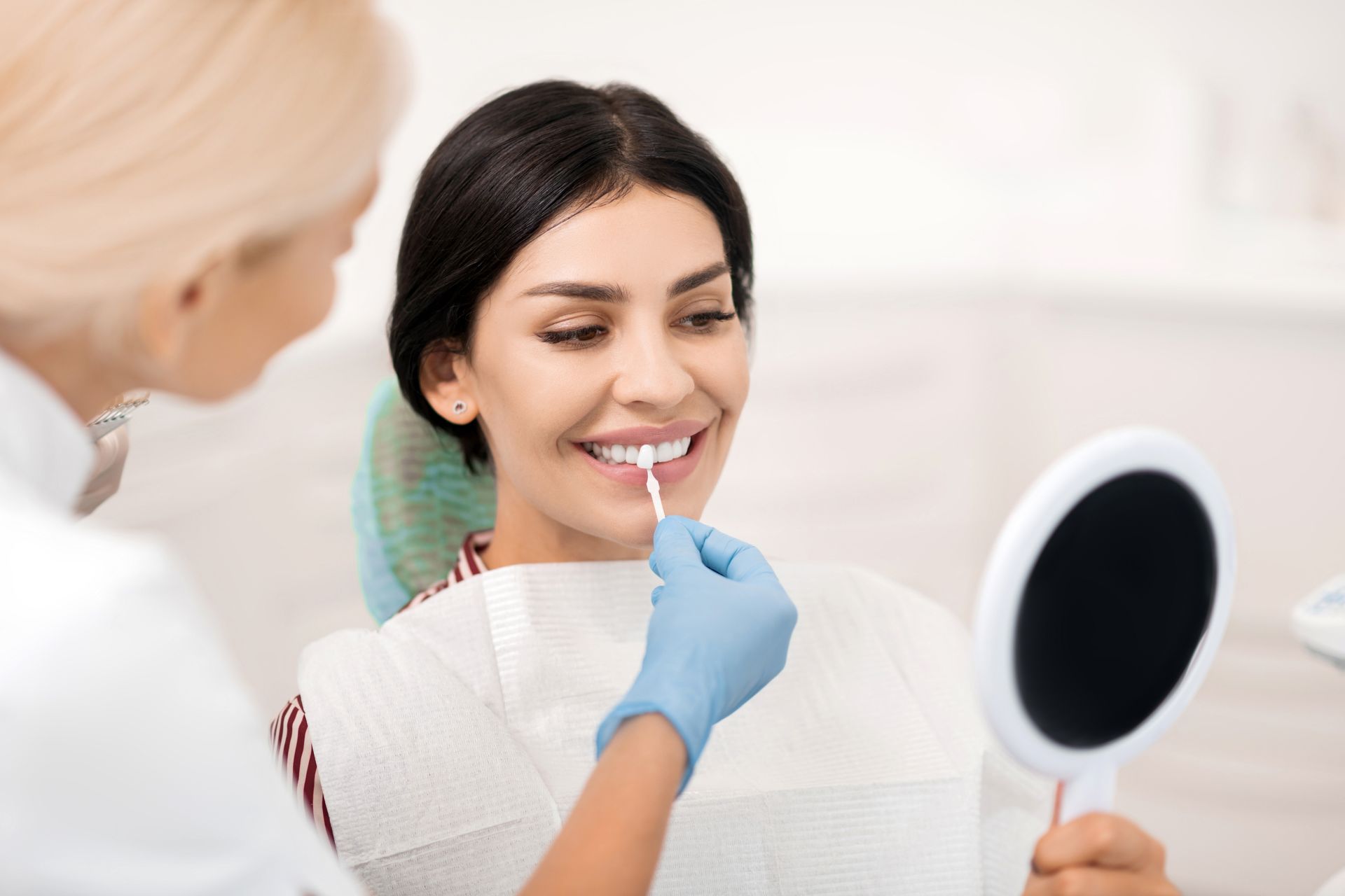Dentist applying lipstick to smiling woman holding a mirror in a bright clinic