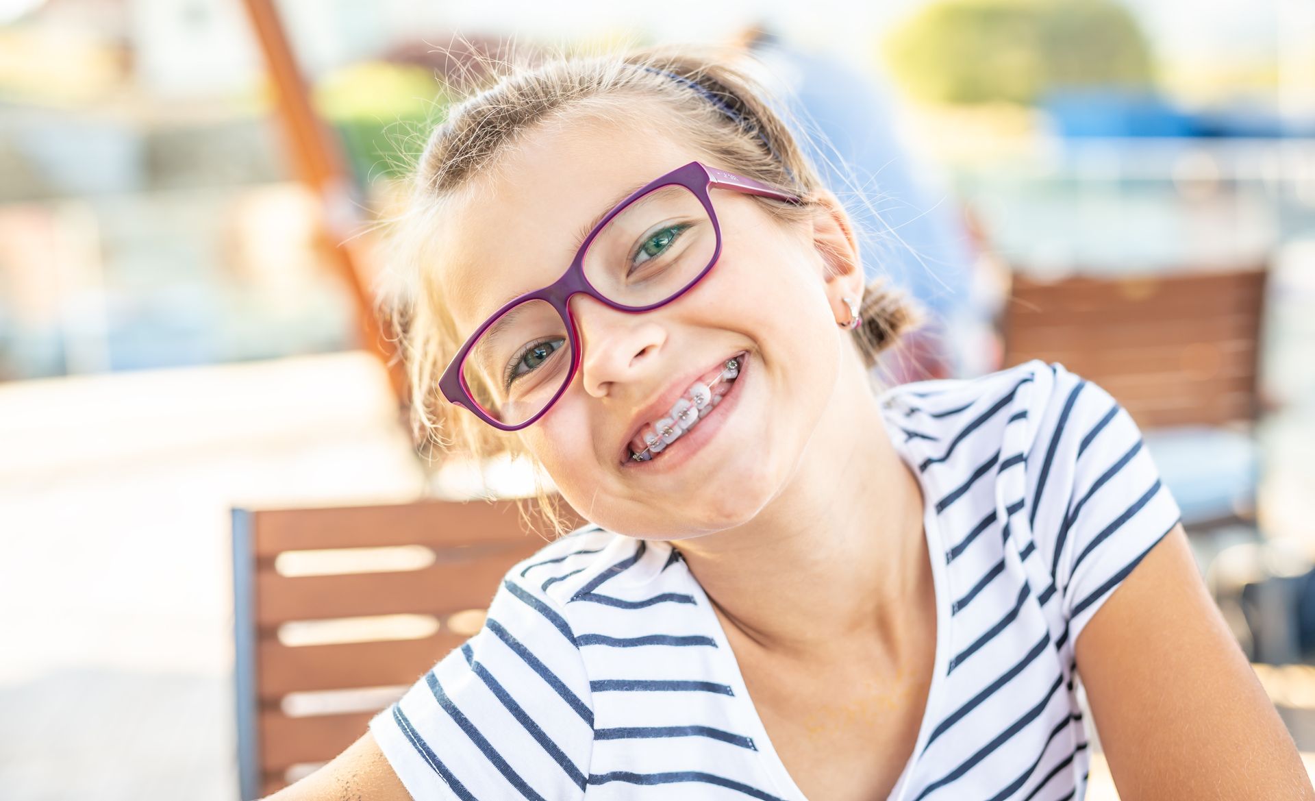 Smiling child wearing purple glasses and a striped shirt, leaning forward outdoors in bright sunlight