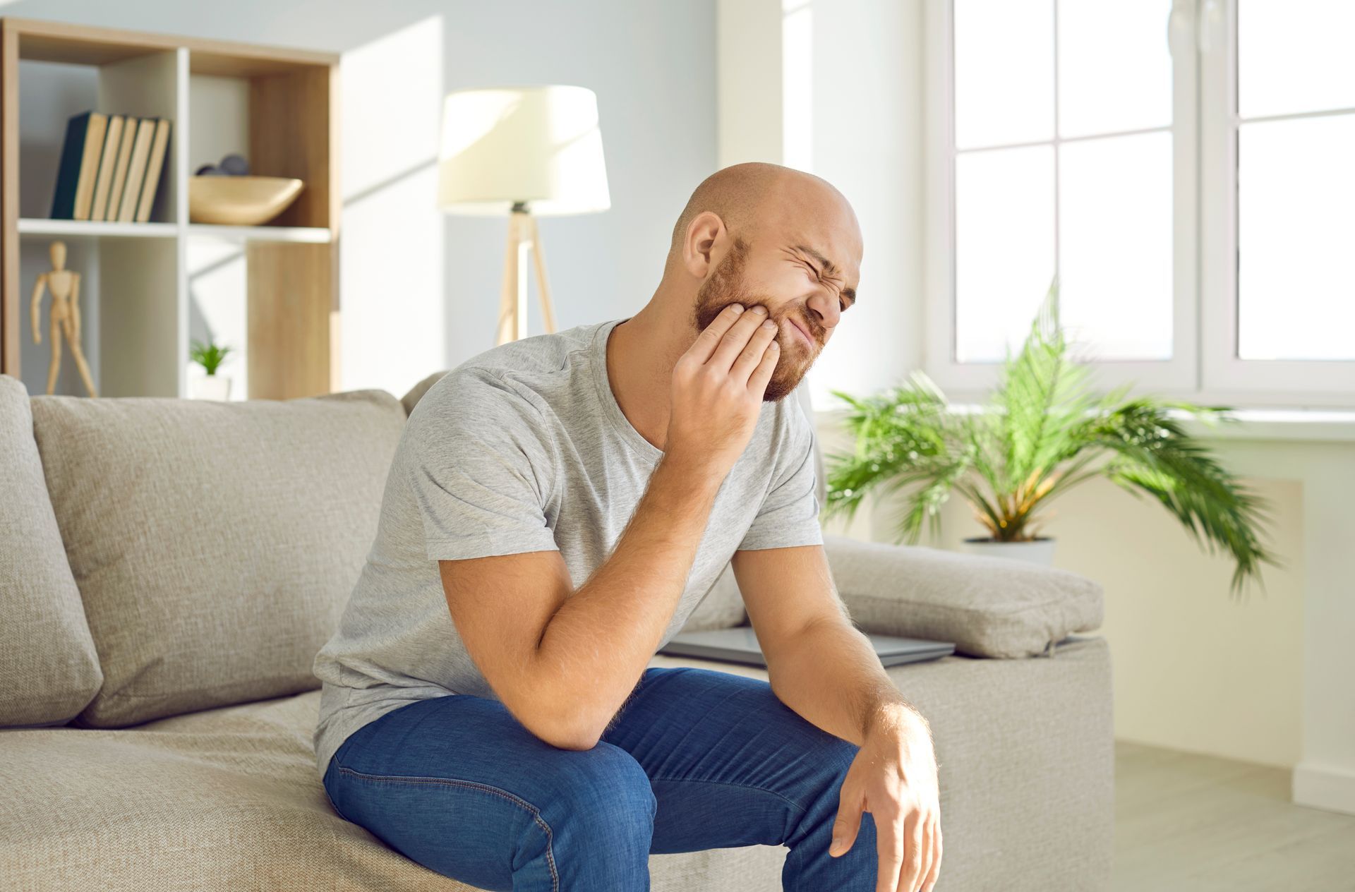 Person sitting on a couch, holding their cheek with a pained expression in a bright living room.