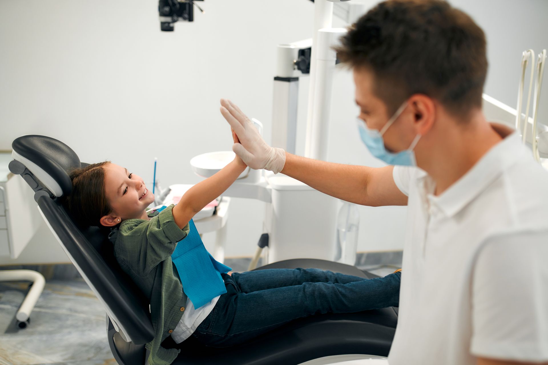 Dentist high-fiving a child patient in a dental chair, with both wearing masks in a bright clinic.