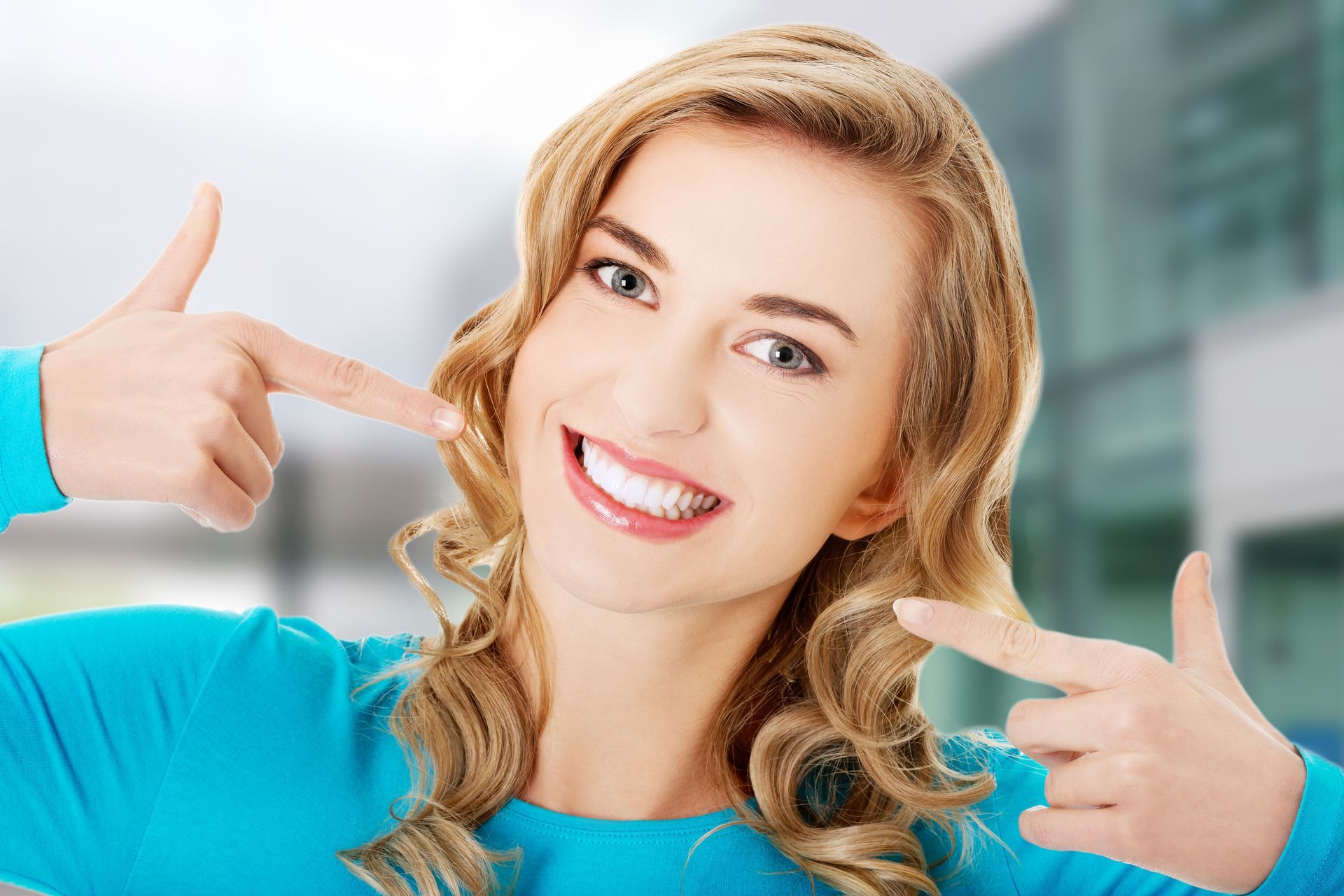 Smiling person in a blue top pointing at their cheeks in a bright indoor setting