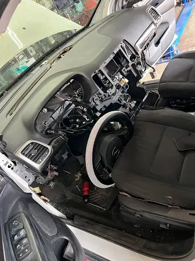 Car interior with dashboard removed, exposing wiring and controls, black seats visible.