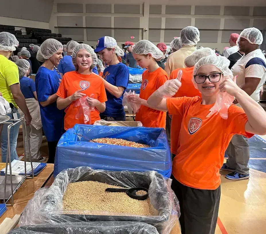 Volunteers in hair nets sorting food at long tables; one person in orange gives a thumbs-up.