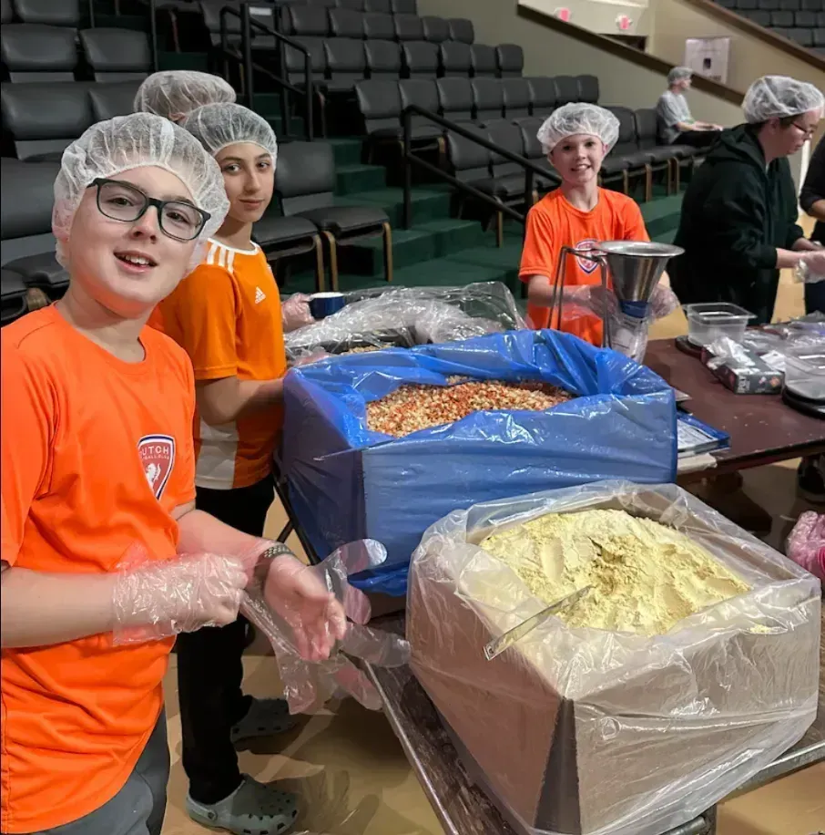 Volunteers in hairnets and orange shirts packaging food at a table in a gymnasium