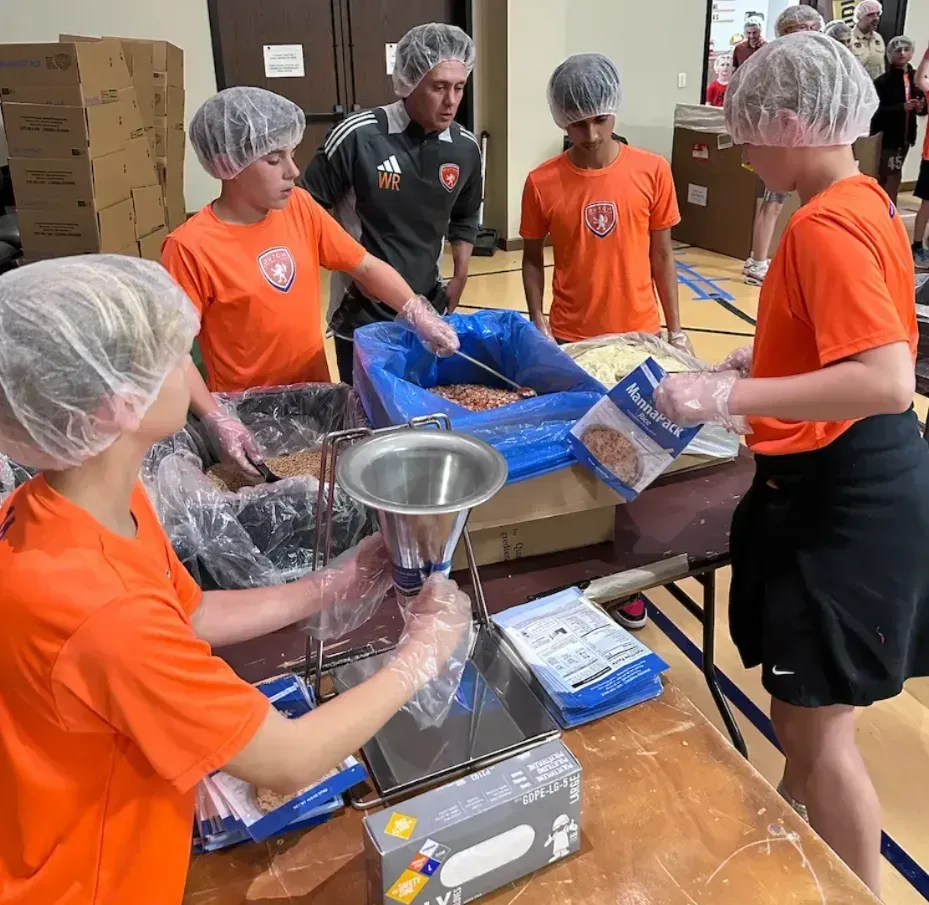 Volunteers in orange shirts bag food at a table in a gymnasium.